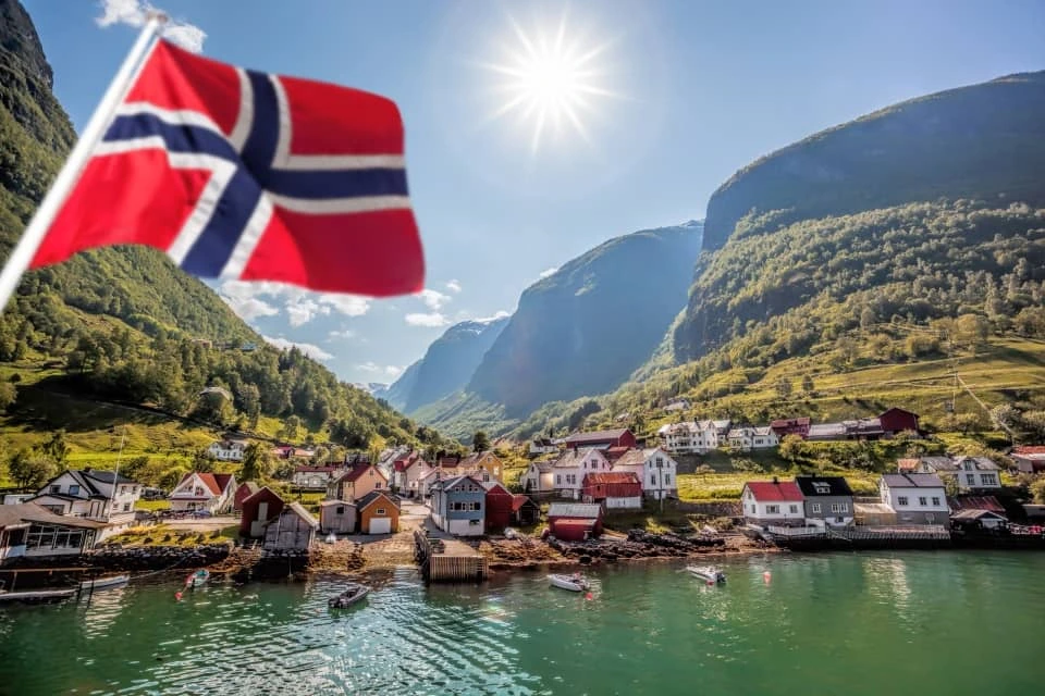 Natural Pharmaceuticals Vistan™ Norra rahvuslipp lehvib fjordi kalda ääres asuva väikese küla taustal päikesepaistelises mägimaastikus.